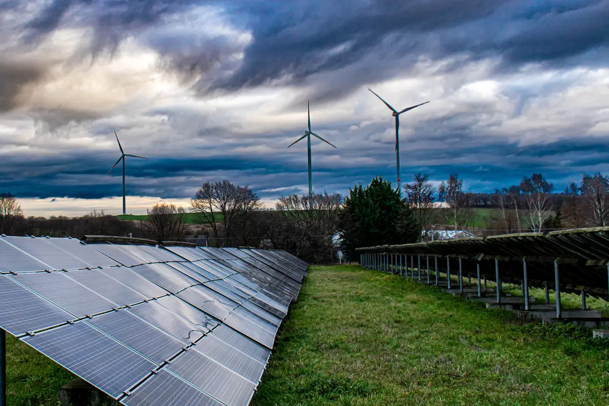 A landscape featuring rows of solar panels and tall wind turbines under a dramatic sky, representing green energy expansion.