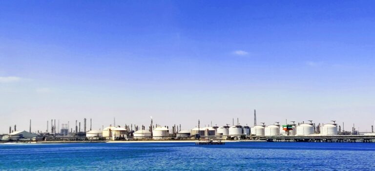 A wide-angle view of white industrial oil storage tanks and refinery infrastructure along a coastline under a clear blue sky.